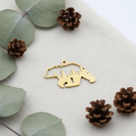 Gold bear-shaped pendant with mountain design held between fingers against a blurred background